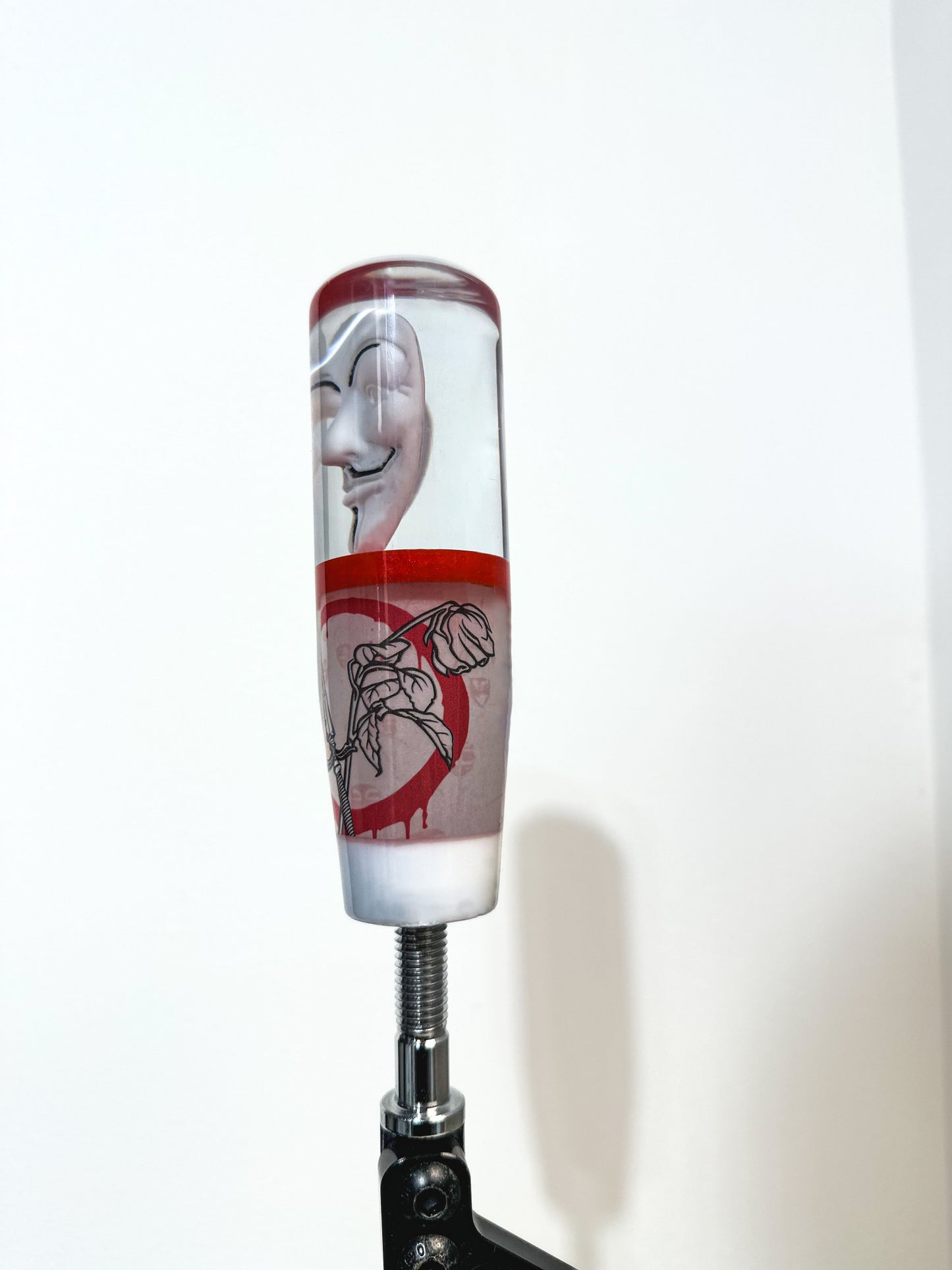 Guy Fawkes mask shift knob made by nice knob, bro. an American shift knob company. Shifter transitions from Fawkes mask to red layer, followed by v for vendetta inspired design. dead rose crossed with dagger on bloody circle. in background a legion of anonymous. final layer white. great universal stick shifter weighted for cars. 